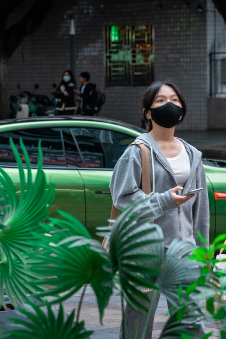 Woman in a gray hoodie and black mask holding a phone with a green car in the background