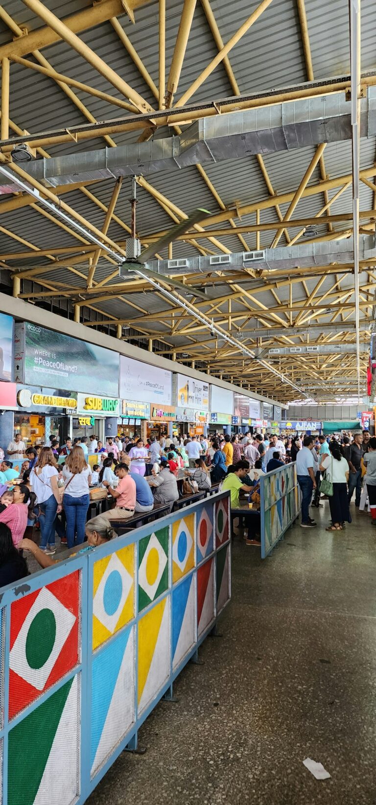 A busy food court area filled with people seated and standing near multiple food stalls.