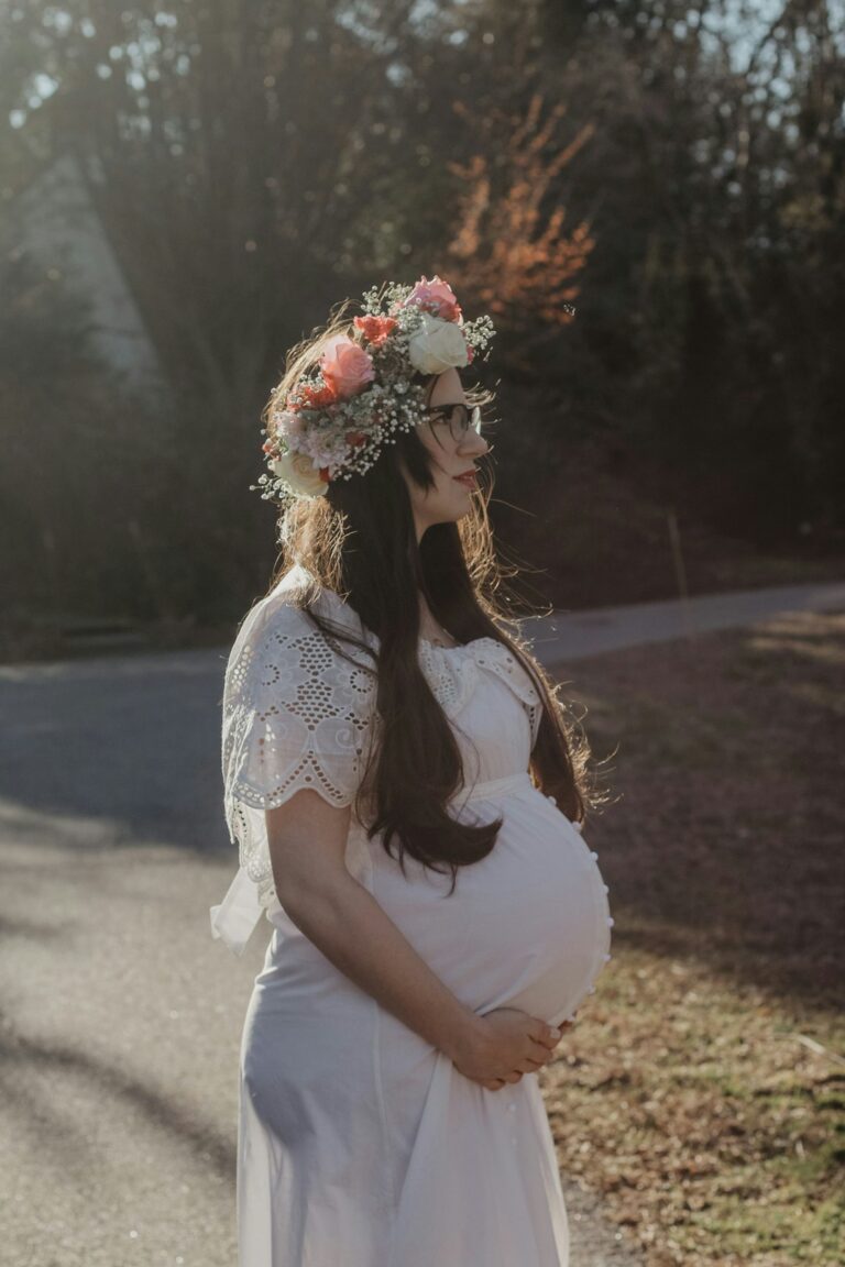 Pregnant woman in white dress and floral crown standing outdoors in peaceful light