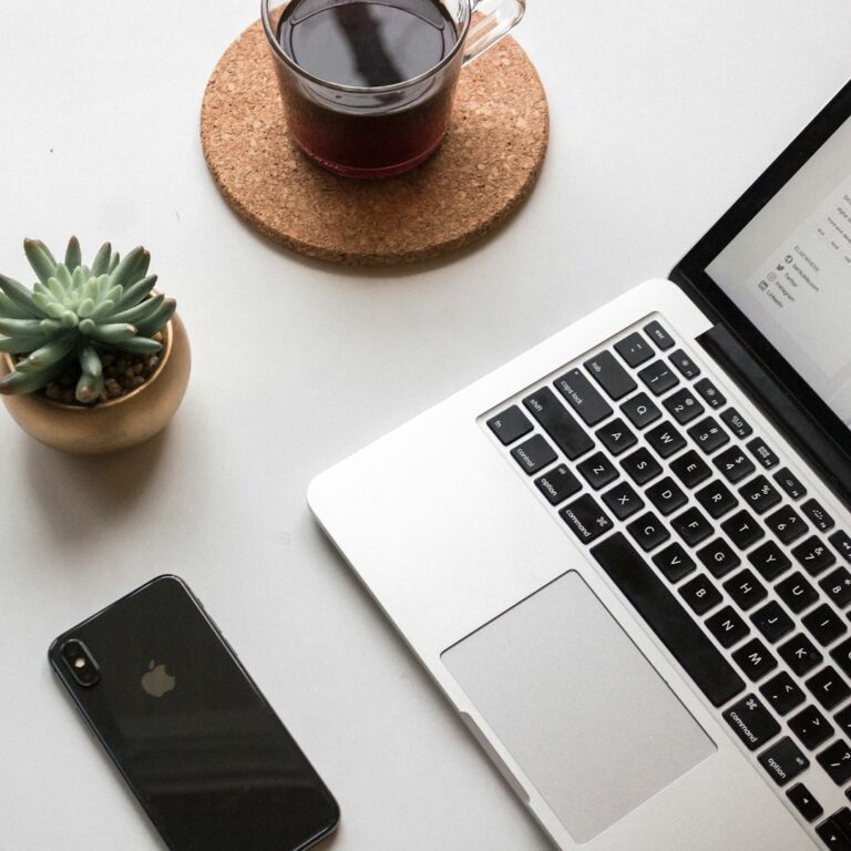 Top view of a workspace with a MacBook, iPhone, cup of coffee on a cork coaster, and a small potted succulent on a white desk.