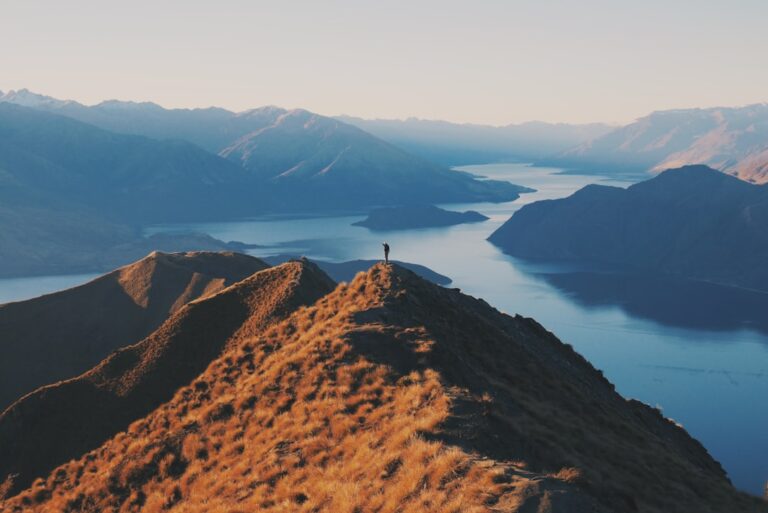 A lone person stands on a mountain peak overlooking lakes and mountains at sunrise.