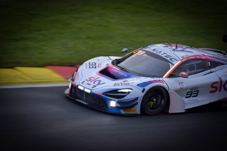 Close-up of a racing car with number 93, displaying the Sky logo, and adorned with multiple sponsors as it speeds on a race track under artificial lights.