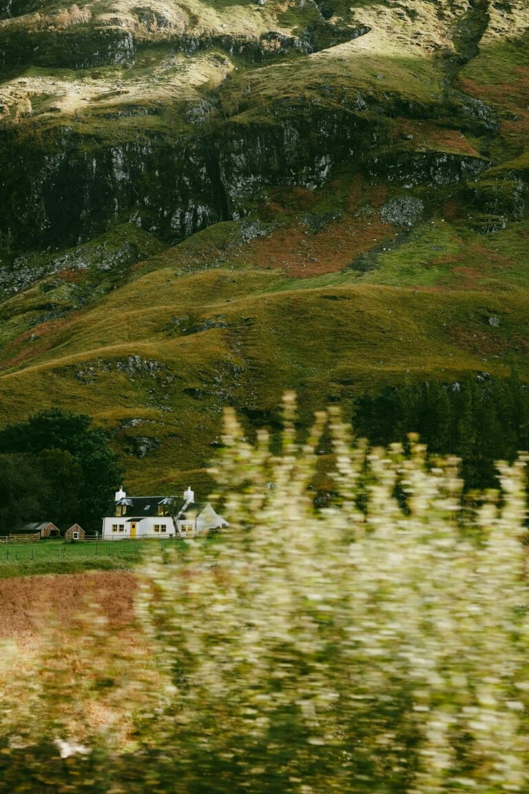 A quaint white cottage with yellow windows situated in a lush, green valley surrounded by hills.