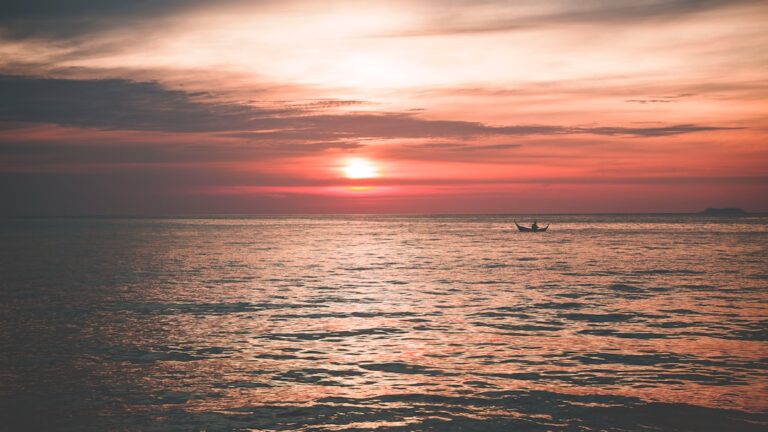 Calm ocean at sunset with a small boat in the distance, orange and pink hues filling the sky.