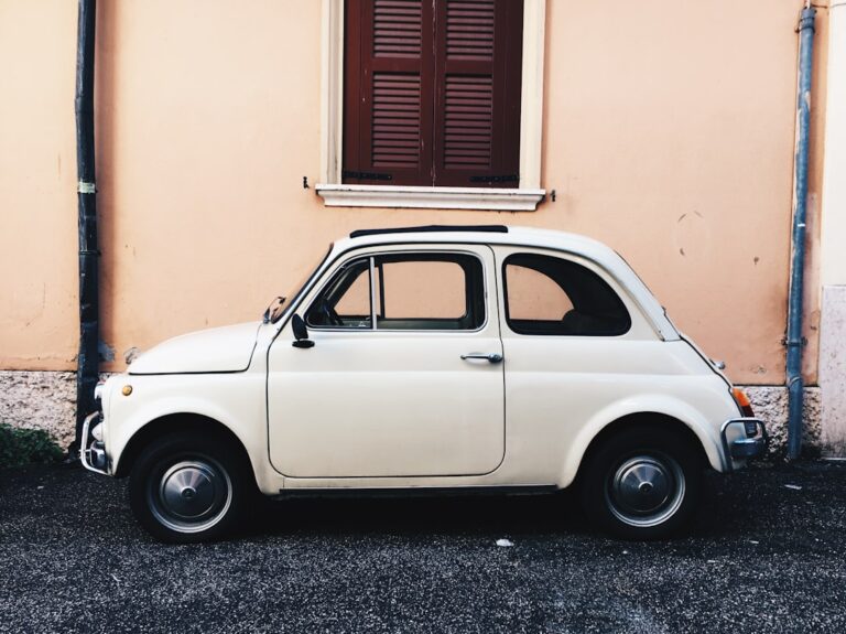Side view of a classic white vintage car parked in front of a beige wall with a shuttered window.