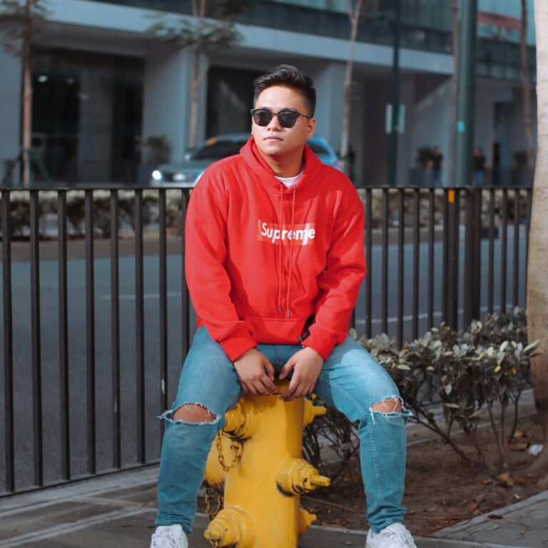 A person in a red Supreme hoodie and ripped jeans sitting on a yellow fire hydrant on a city street.