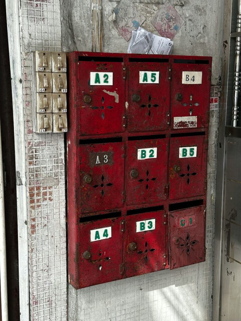Eight rustic red metal mailboxes with peeling paint, labeled with alphanumeric codes, mounted on a tiled wall next to a row of electrical switches.
