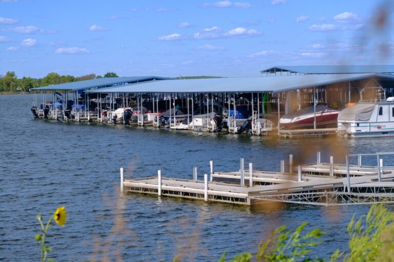 Several boats are docked at a covered marina on a sunny day with blue sky and fluffy clouds.