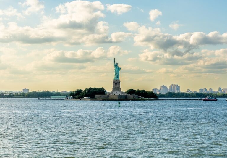 A view of the Statue of Liberty from across the water with buildings in the background and a cloudy sky.