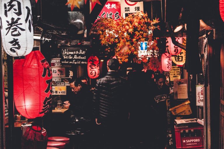 People enjoying food and drinks in a bustling Japanese izakaya alley with traditional lanterns and signs at night