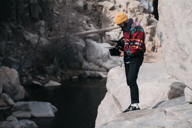 Person standing near a rocky stream taking photos with a professional camera setup, wearing a colorful jacket and yellow beanie.