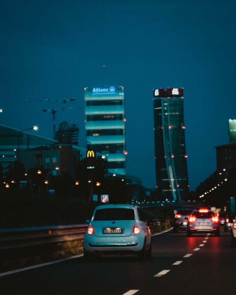 Cars driving at night in a city with high-rise buildings illuminated in the background.